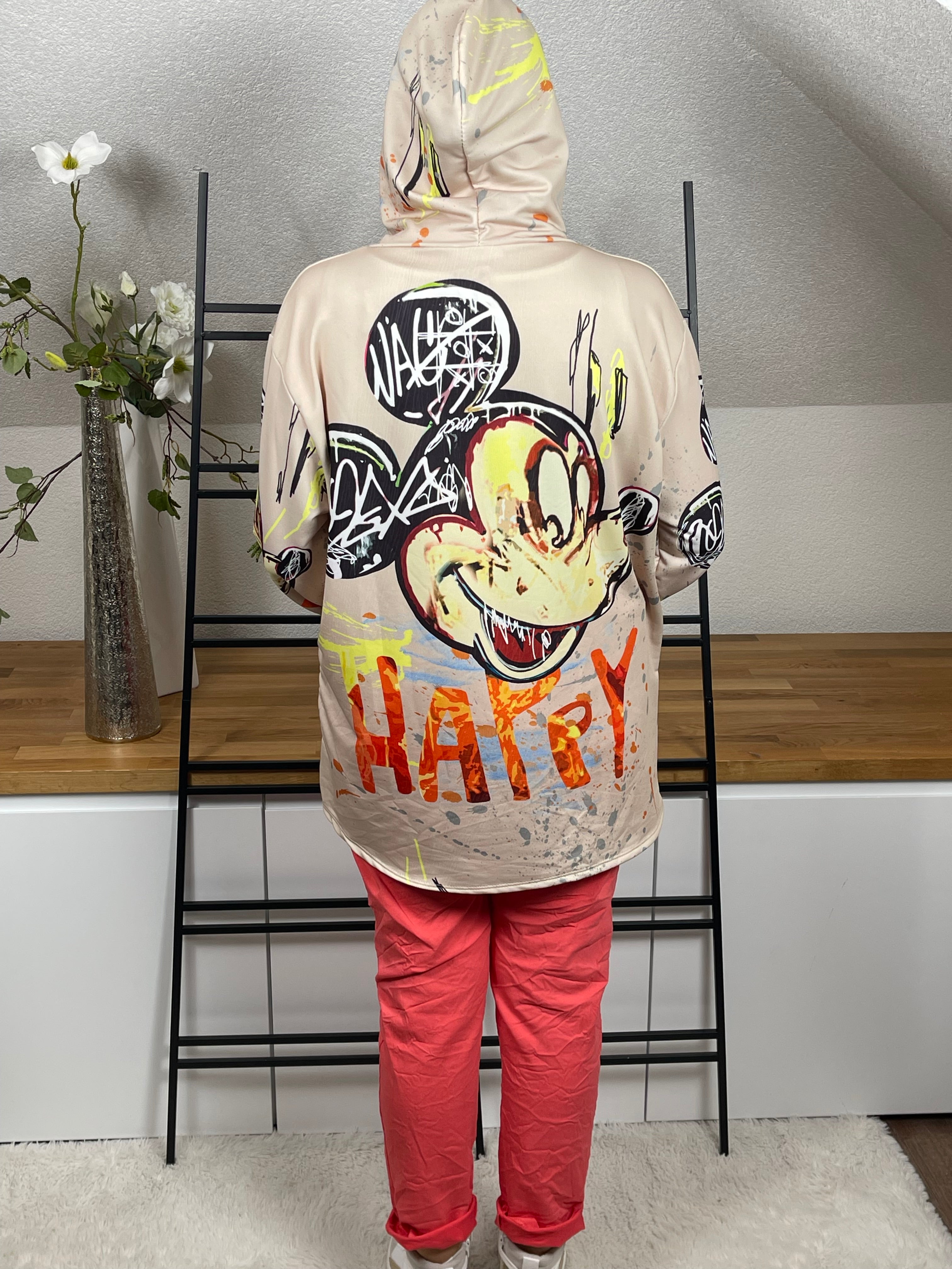 Oversize-Hoodie in beige, mit Mickey - Print von hinten, zeigt die locker fallende Passform und die stylische Kapuze. Kombiniert mit einer korallfarbenen Jogpant und weiß - goldenen High - Sneakern für einen modernen Look.