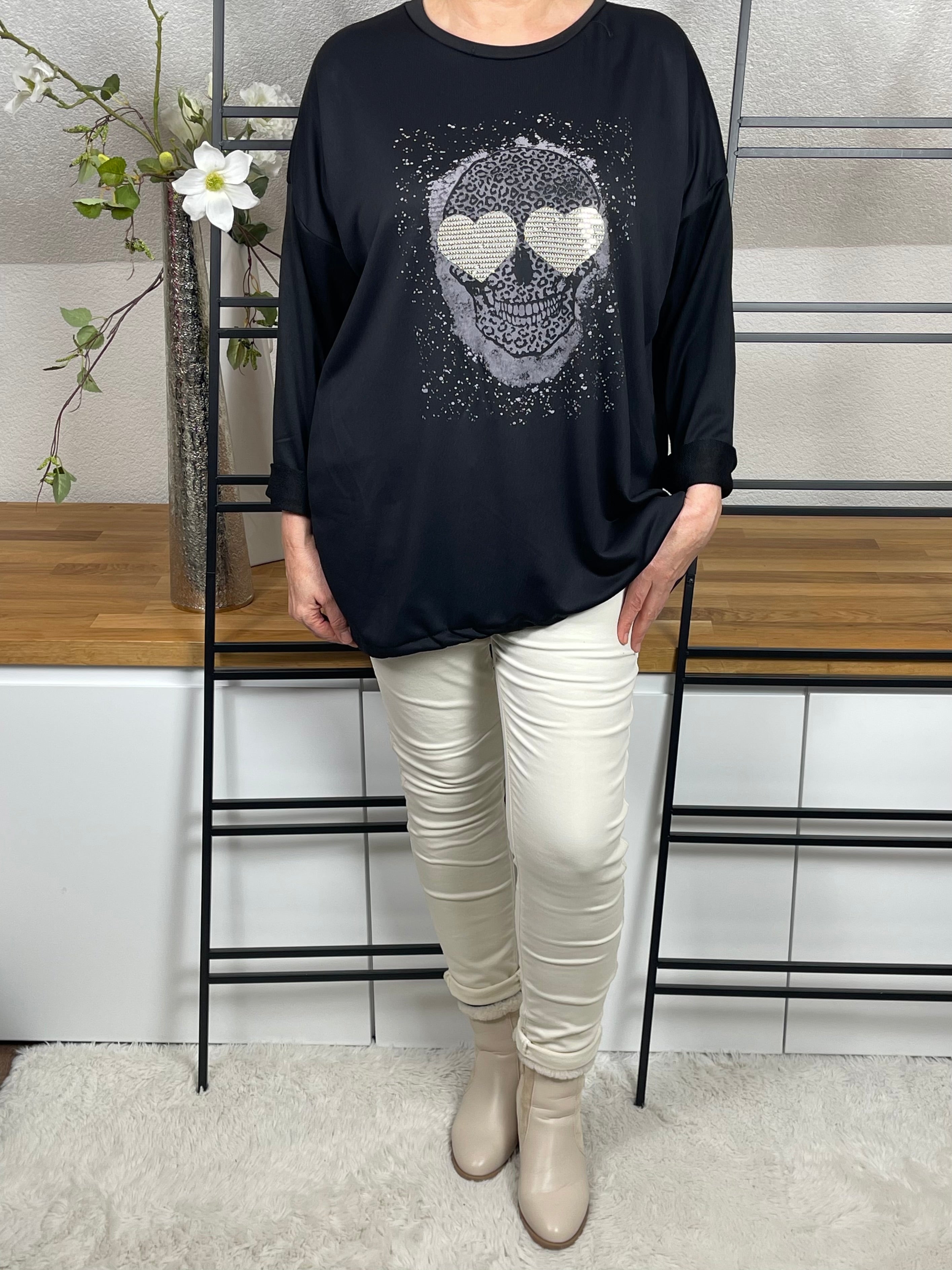 Vorderansicht des schwarzen  Oversize-Damen-Pullovers  mit Totenkopf-Print, Glitzer-Details, locker getragen mit einer cremeweißen Jogpant und cremeweißen Stiefeletten