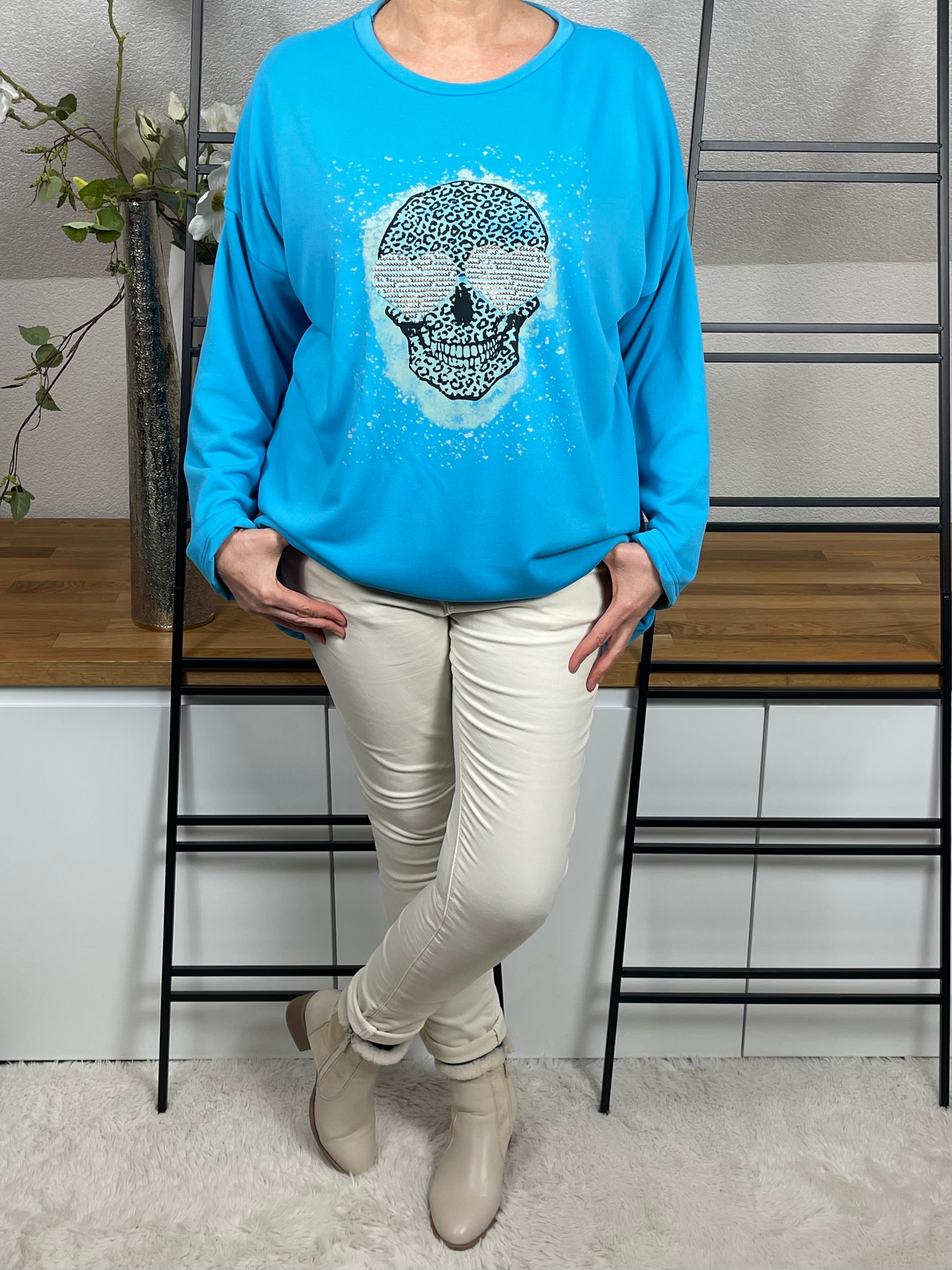 Vorderansicht des türkisen Oversize-Damen-Pullovers  mit Totenkopf-Print, Glitzer-Details, Länge reguliert mit Kordel, getragen mit einer cremeweißen Jogpant und cremeweißen Stiefeletten