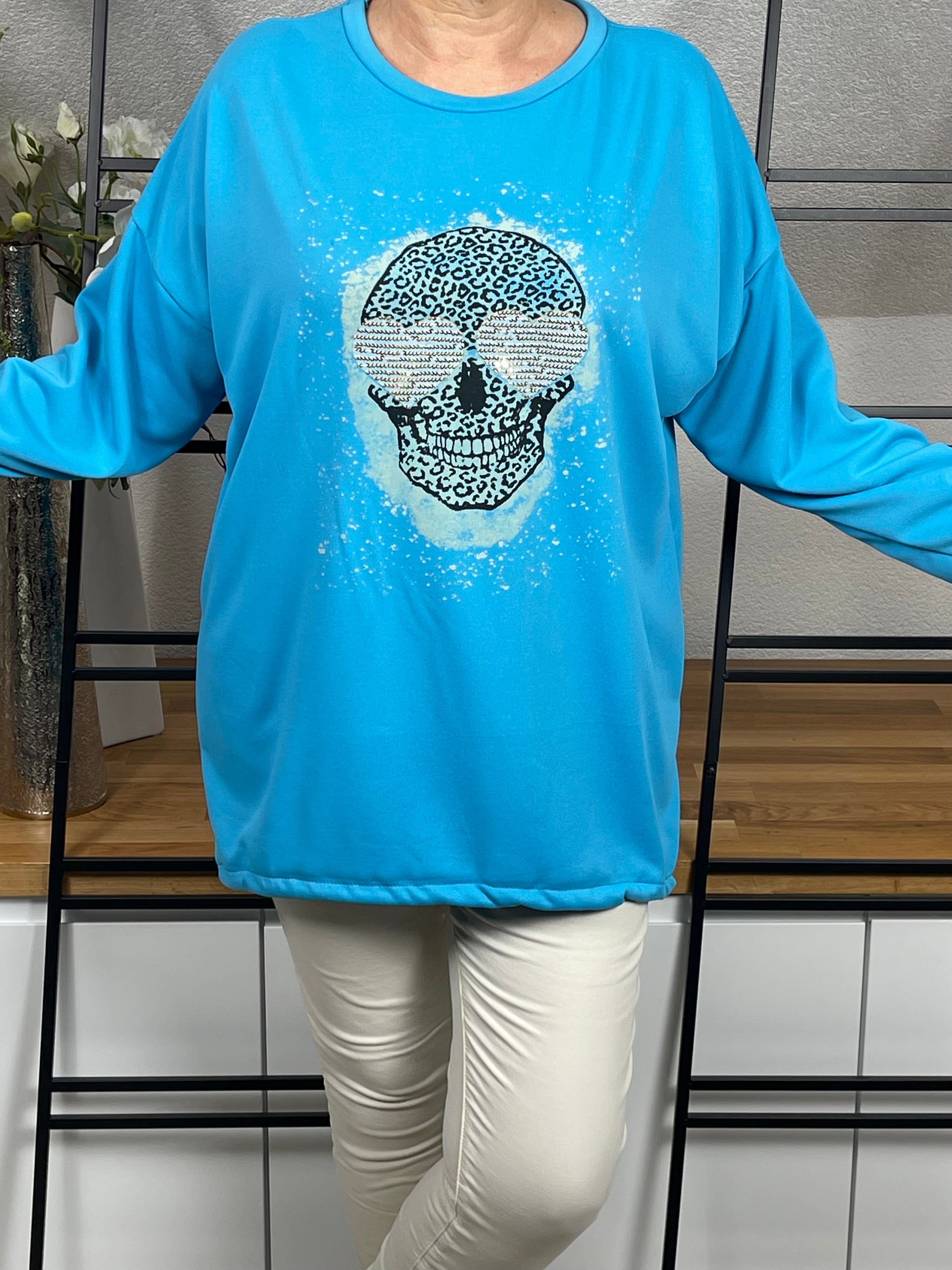 Vorderansicht des türkisen Oversize-Damen-Pullovers mit Totenkopf-Print, Glitzer-Details, in ganzer Länge mit ausgestreckten Armen
