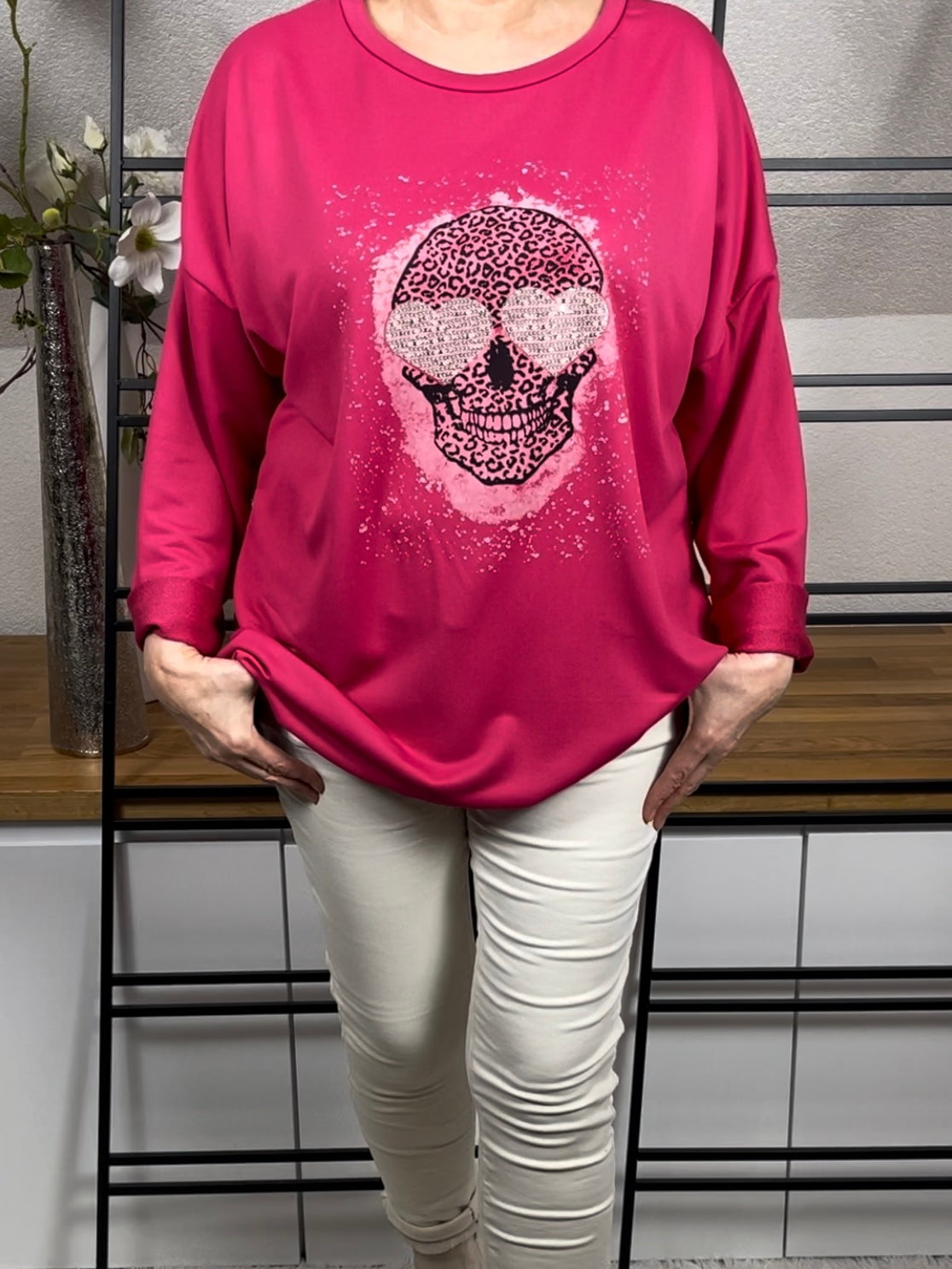 Vorderansicht des pinkfarbenen Oversize-Damen-Pullovers  mit Totenkopf-Print, Glitzer-Details, Länge reguliert mit Kordel, getragen mit einer cremeweißen Jogpant und cremeweißen Stiefeletten