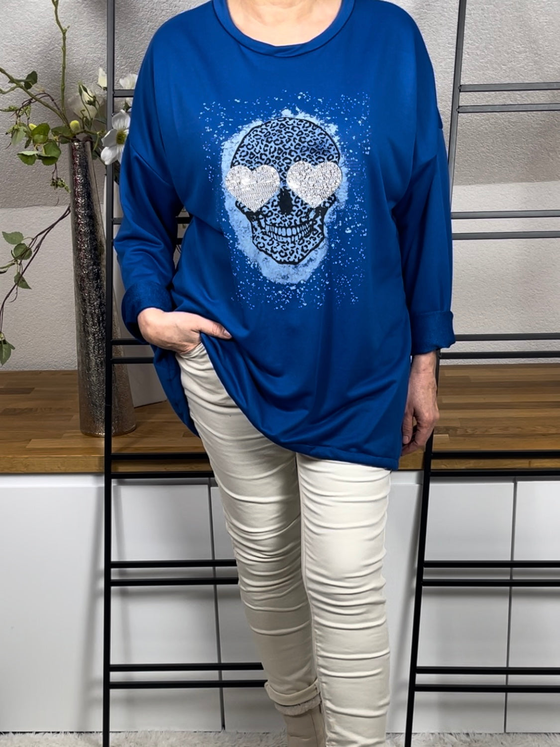 Vorderansicht des azurblauen  Oversize-Damen-Pullovers  mit Totenkopf-Print, Glitzer-Details, lässig getragen mit einer cremeweißen Jogpant und cremeweißen Stiefeletten