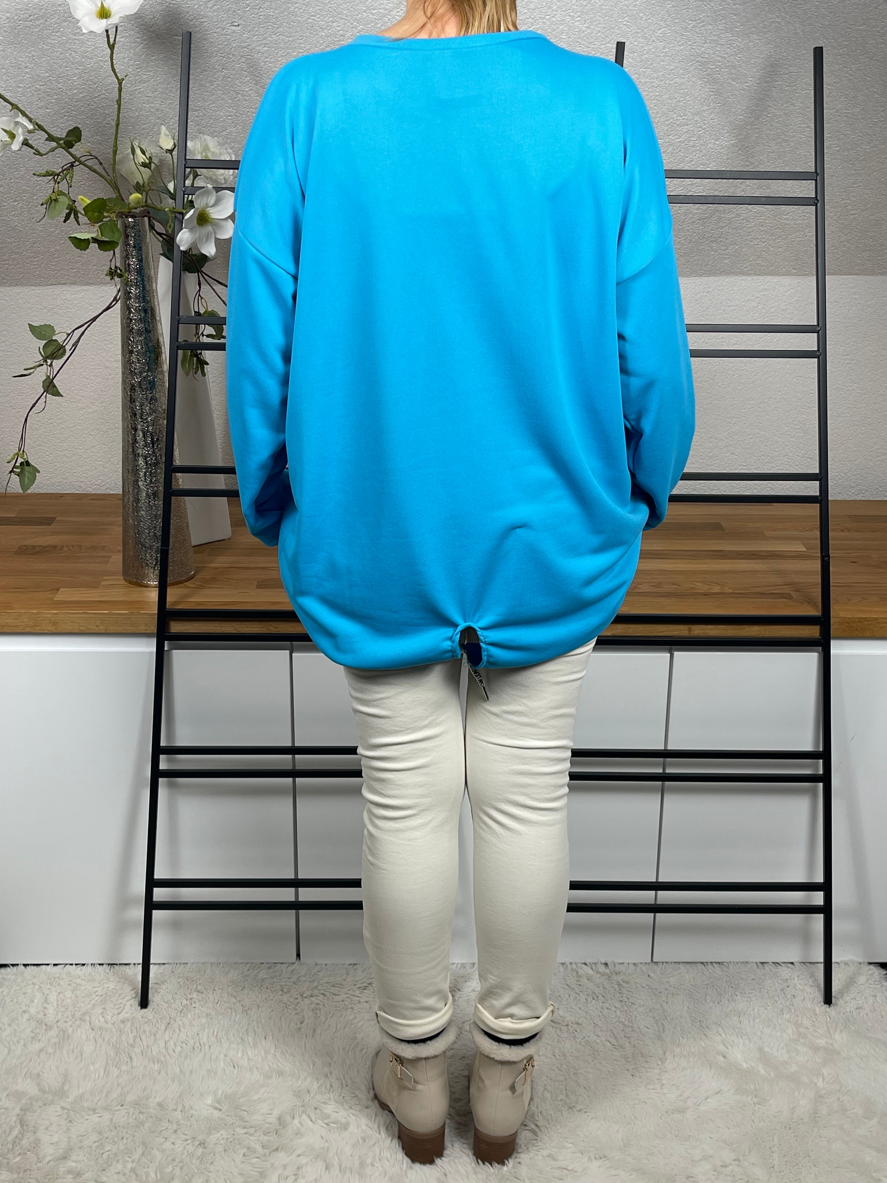 Rückenansicht des türkisen Oversize-Damen-Pullovers  mit Totenkopf-Print, Glitzer-Details, Länge reguliert mit Kordel, getragen mit einer cremeweißen Jogpant und cremeweißen Stiefeletten